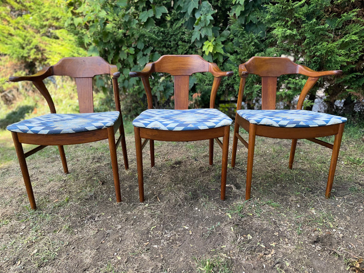 Three wooden armchairs with blue and white patterned seat cushions, arranged side-by-side on a dirt and grass lawn with green foliage in the background.