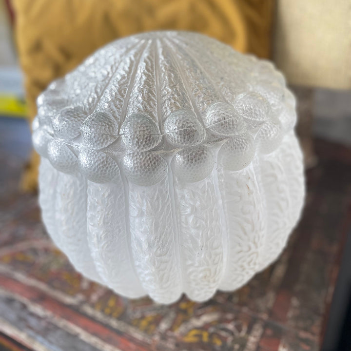 vintage Art Deco style white frosted glass lamp shade with a textured floral pattern, resting on a rustic wooden surface with a yellow pillow in the background.
