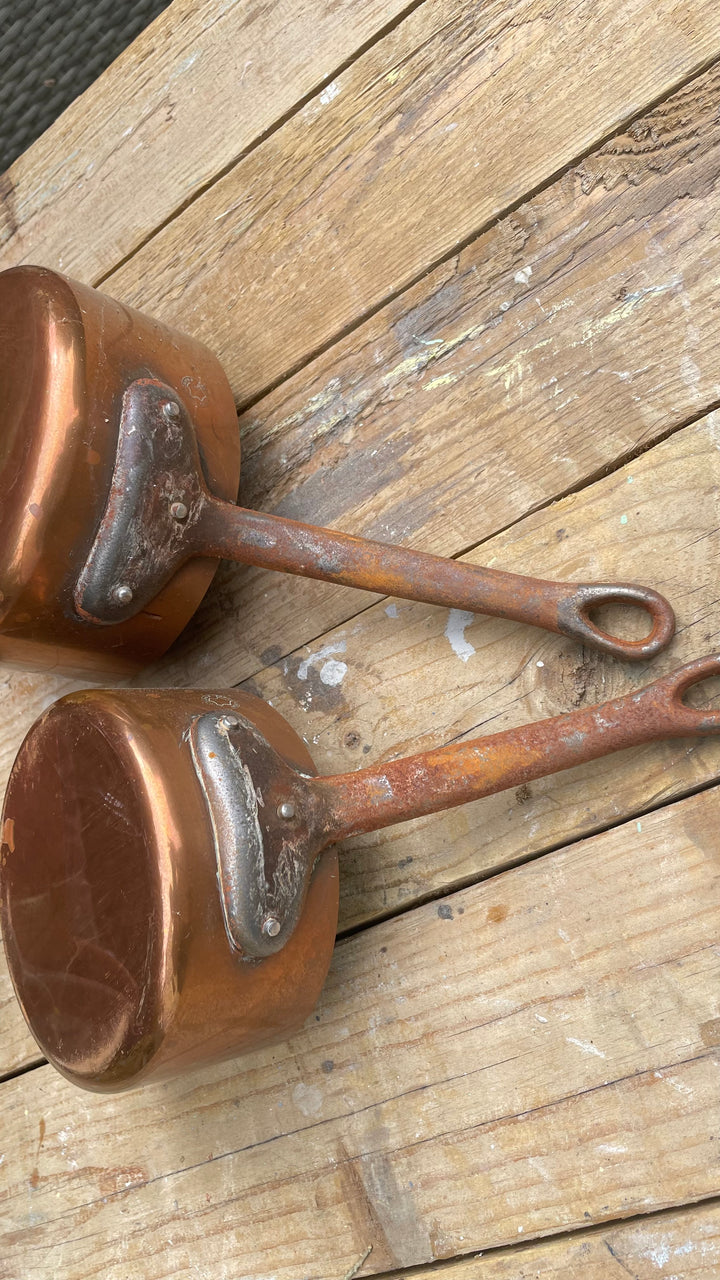 Two vintage copper saucepans with long handles and looped ends, resting on a rustic wooden plank surface.
Details