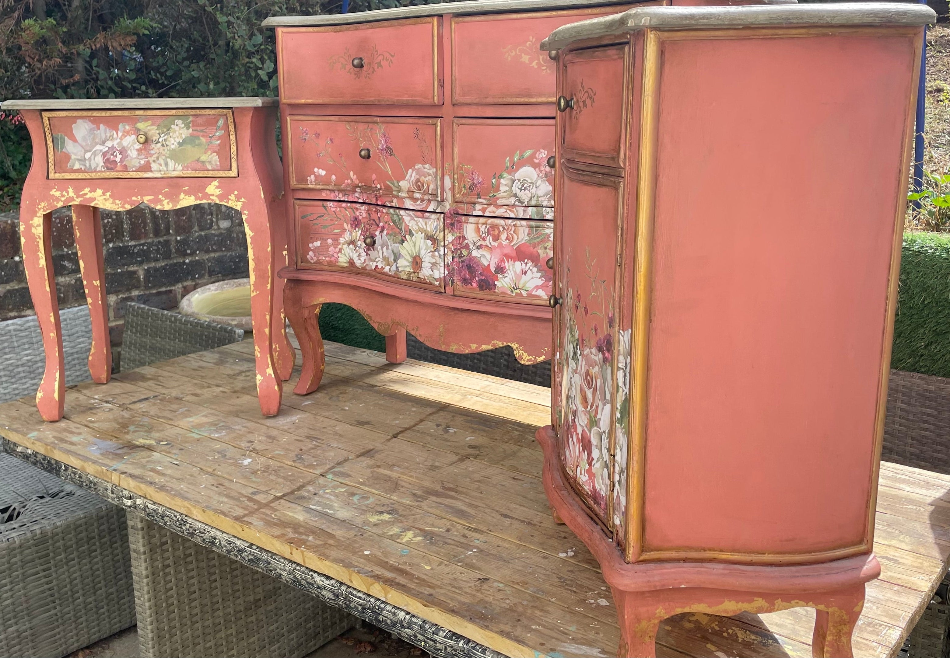 Three painted wooden furniture pieces displayed outdoors on a wooden table: a small side table, a three-drawer chest, and a tall cabinet, all in a distressed coral pink color with floral decals and gold-toned trim.
