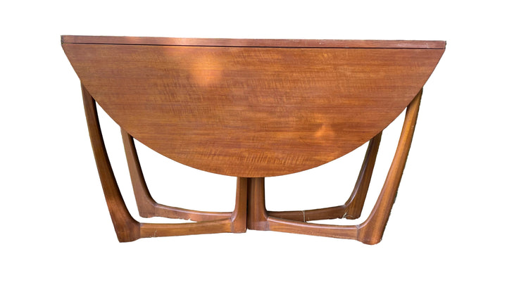 mid-century modern wooden table with a drop-leaf design, featuring a warm reddish-brown wood finish and sculptural curved legs, photographed outdoors on grass with green foliage in the background.