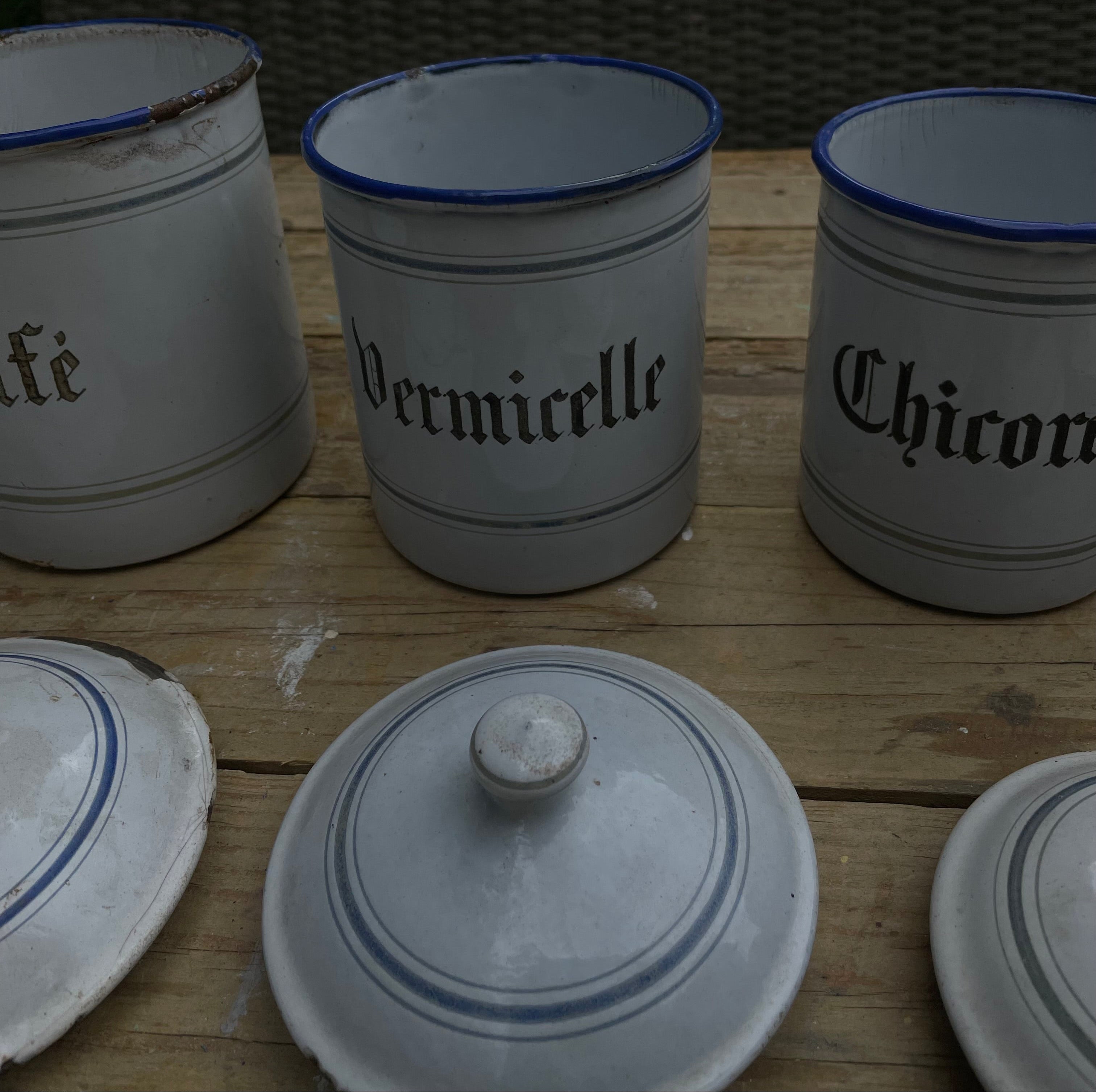 Three white enamel canisters with blue rims and black gothic lettering, labeled 'Vermicelle' and 'Chicory', displayed on a wooden table outdoors.