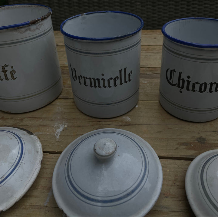 Three white enamel canisters with blue rims and black gothic lettering, labeled 'Vermicelle' and 'Chicory', displayed on a wooden table outdoors.