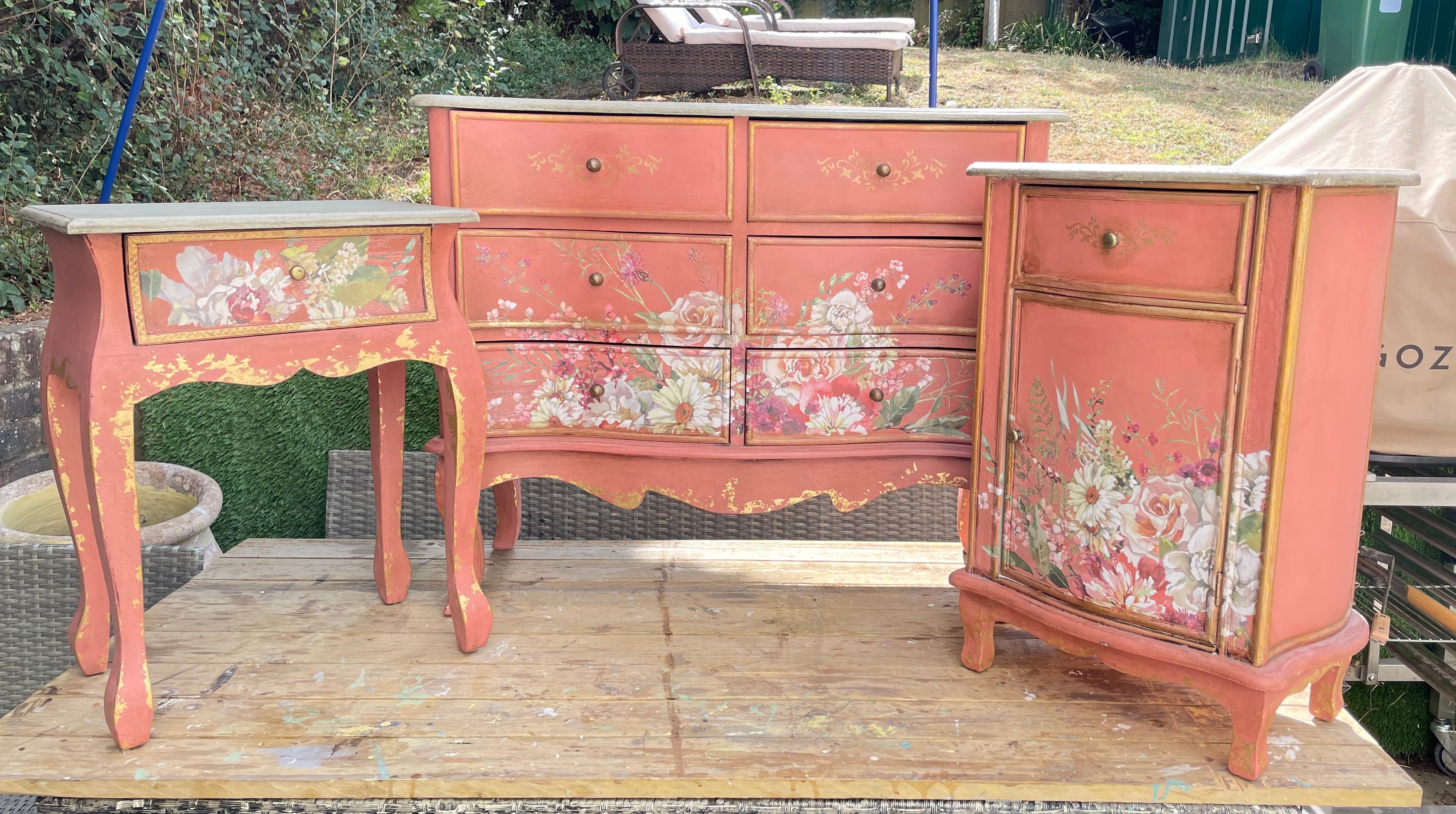 A set of three distressed coral-pink painted wooden furniture pieces with floral decals, displayed on a wooden table in an outdoor backyard setting.