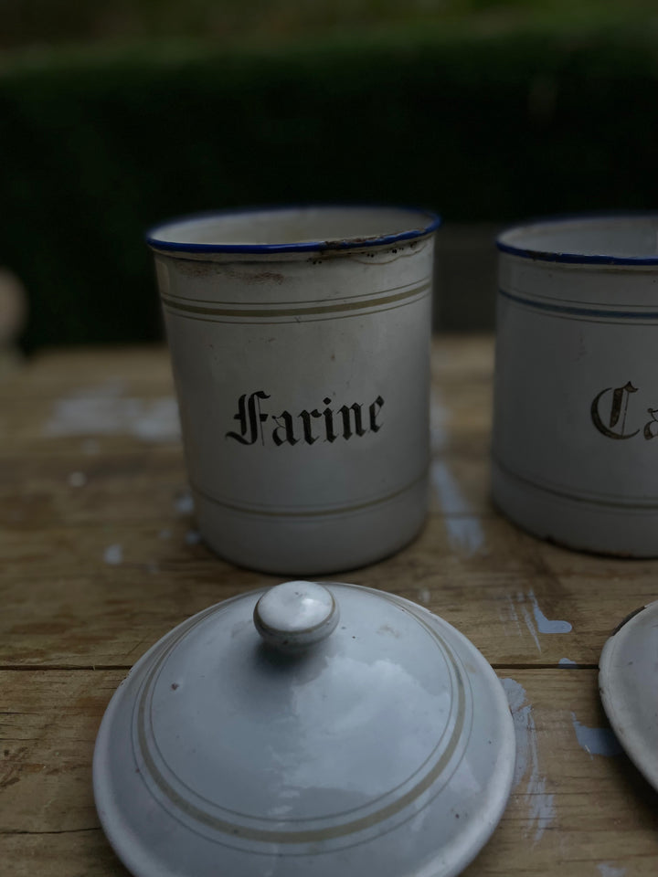 Vintage white enamel canisters with blue rims on a wooden table, featuring one labeled 'Farine' and another partially visible with 'Ca' in gothic lettering, with a matching white lid in the foreground