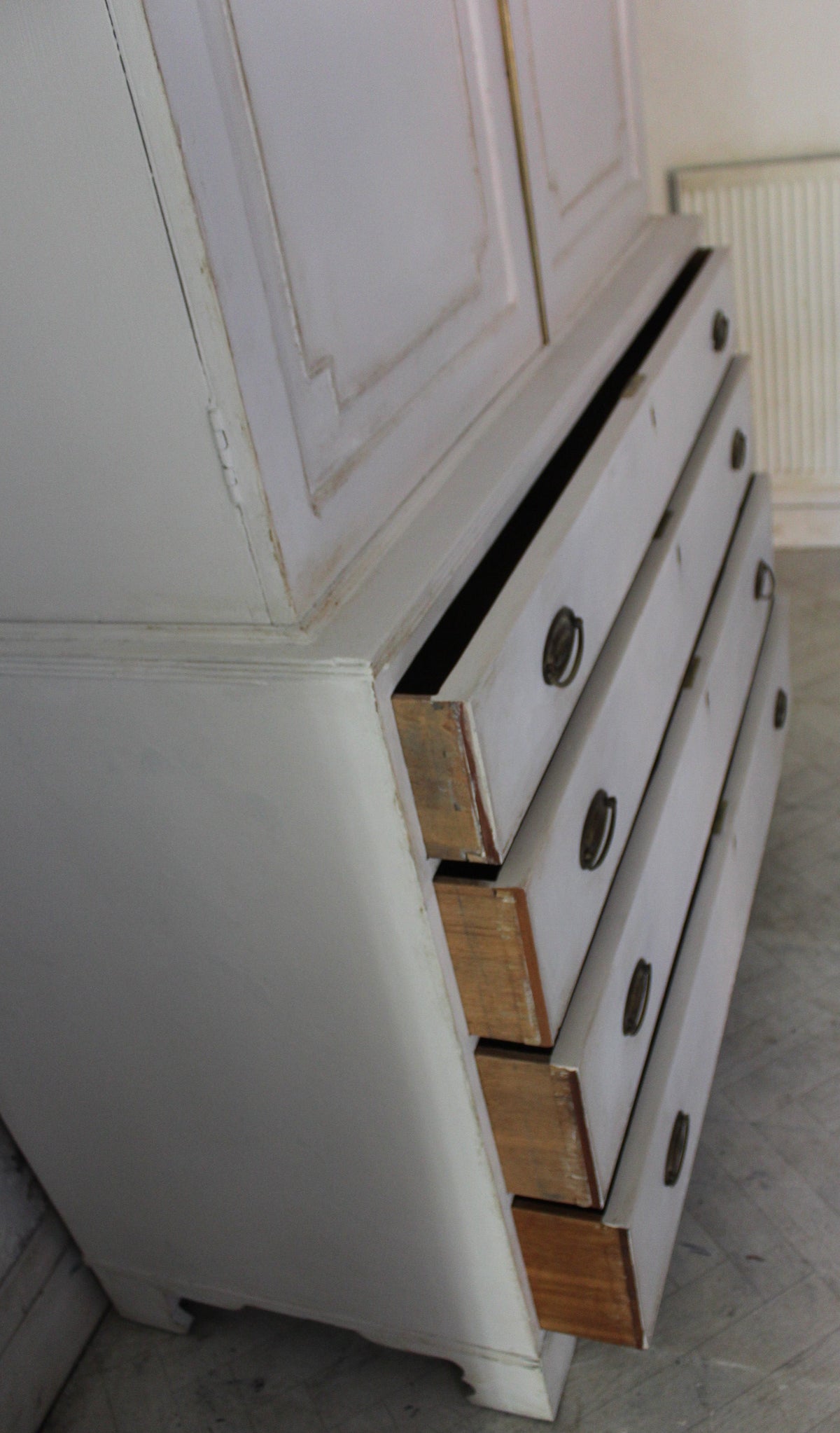 A vintage white painted wooden dresser with several drawers pulled out, showing the natural wood drawer fronts and round dark knobs, set against a room background with a radiator.