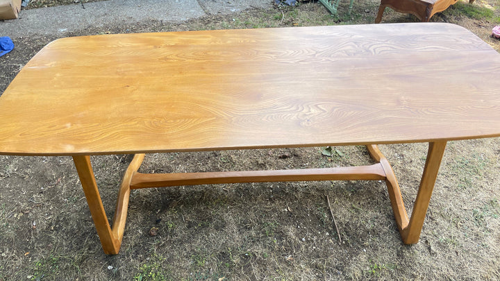 A light brown wooden Elm Ercol dining table with a natural oak grain finish, photographed outdoors on a patch of grass.