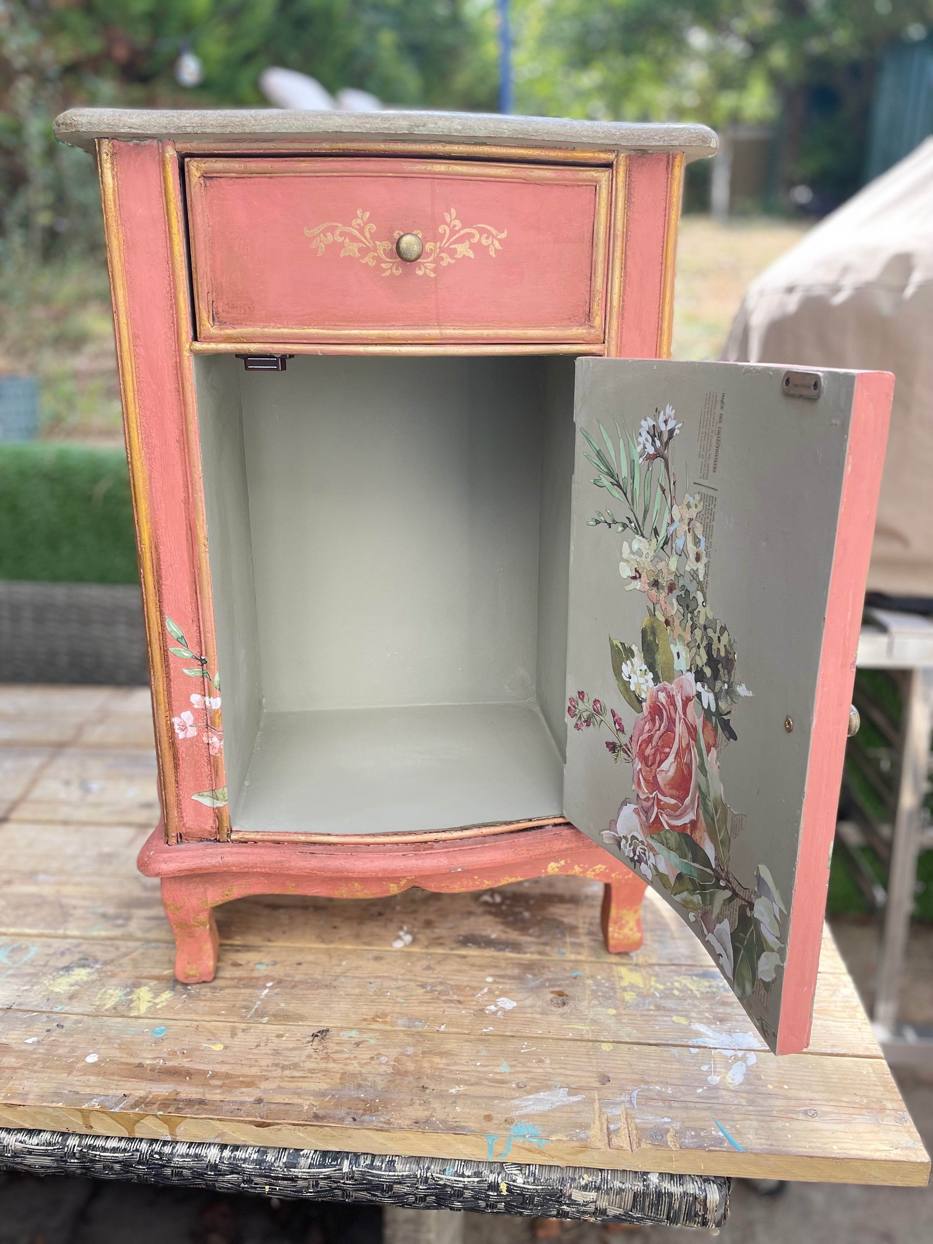 small painted wooden cabinet or side table with a dusty pink/peach finish, gold trim, and floral artwork on the open door, photographed outdoors on a wooden surface with a blurred garden background.