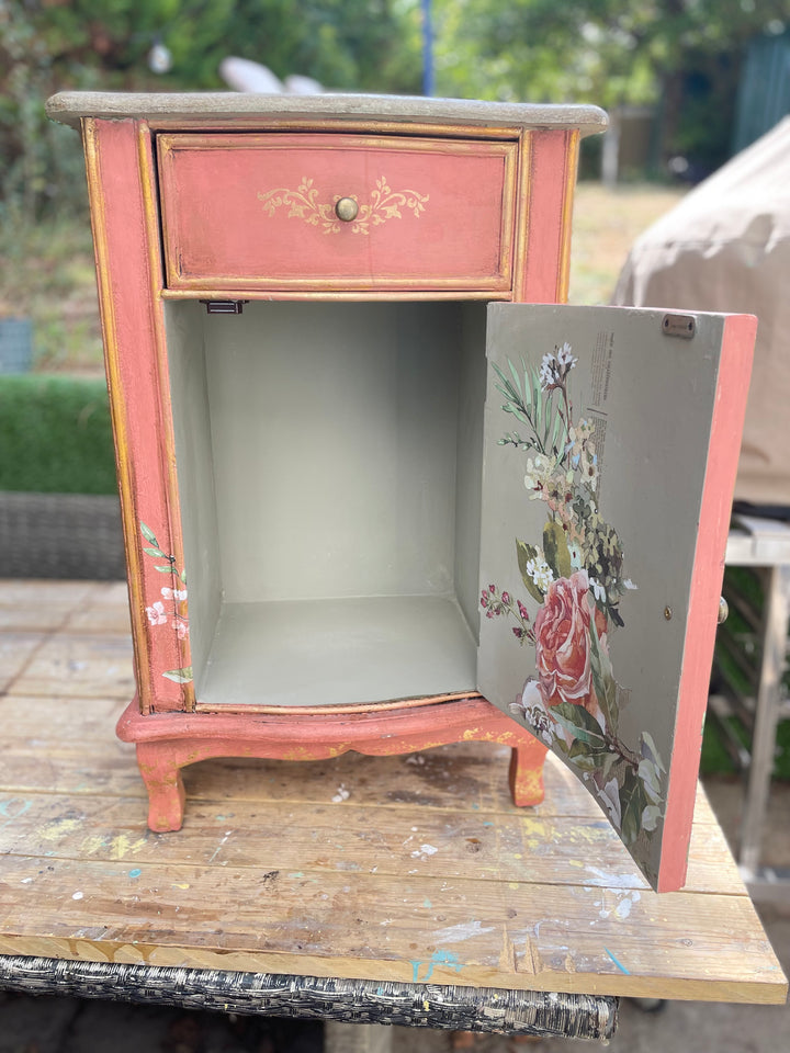 small painted wooden cabinet or side table with a dusty pink/peach finish, gold trim, and floral artwork on the open door, photographed outdoors on a wooden surface with a blurred garden background.