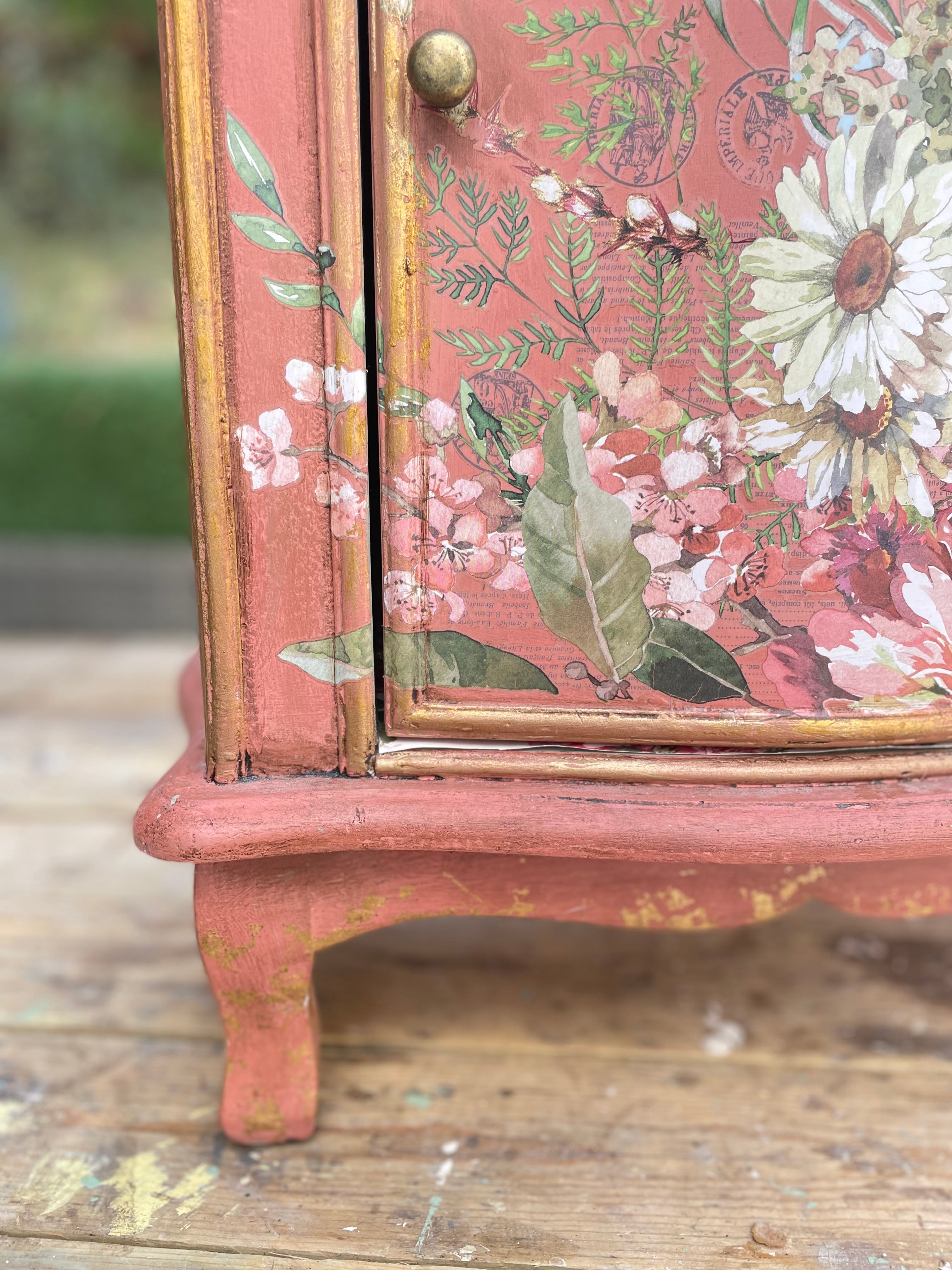 Close-up of a distressed coral-pink cabinet with gold trim and a vintage floral print on the door, resting on a rustic wooden surface.