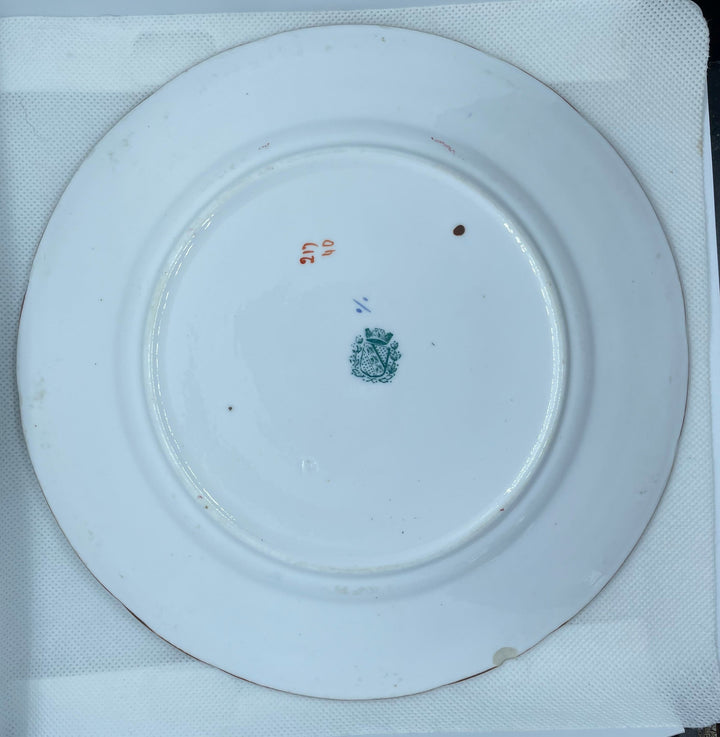 A white ceramic plate photographed from above inside a white protective box lined with paper towel material. The underside of the plate shows a small blue decorative mark and a couple of small orange/red marks.