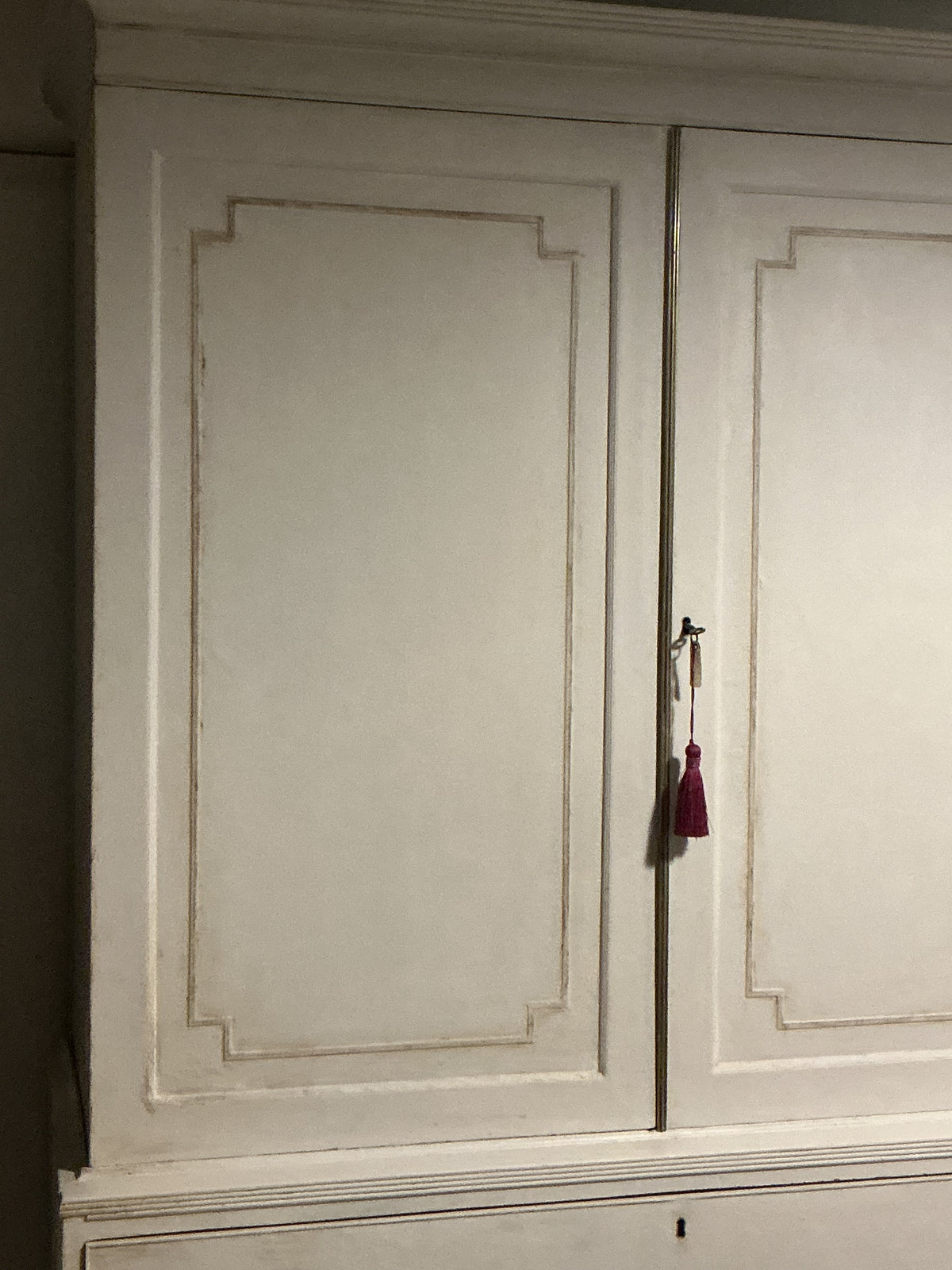A close-up of a distressed white wooden cabinet door with decorative recessed panel lines and a small hanging burgundy tassel keychain