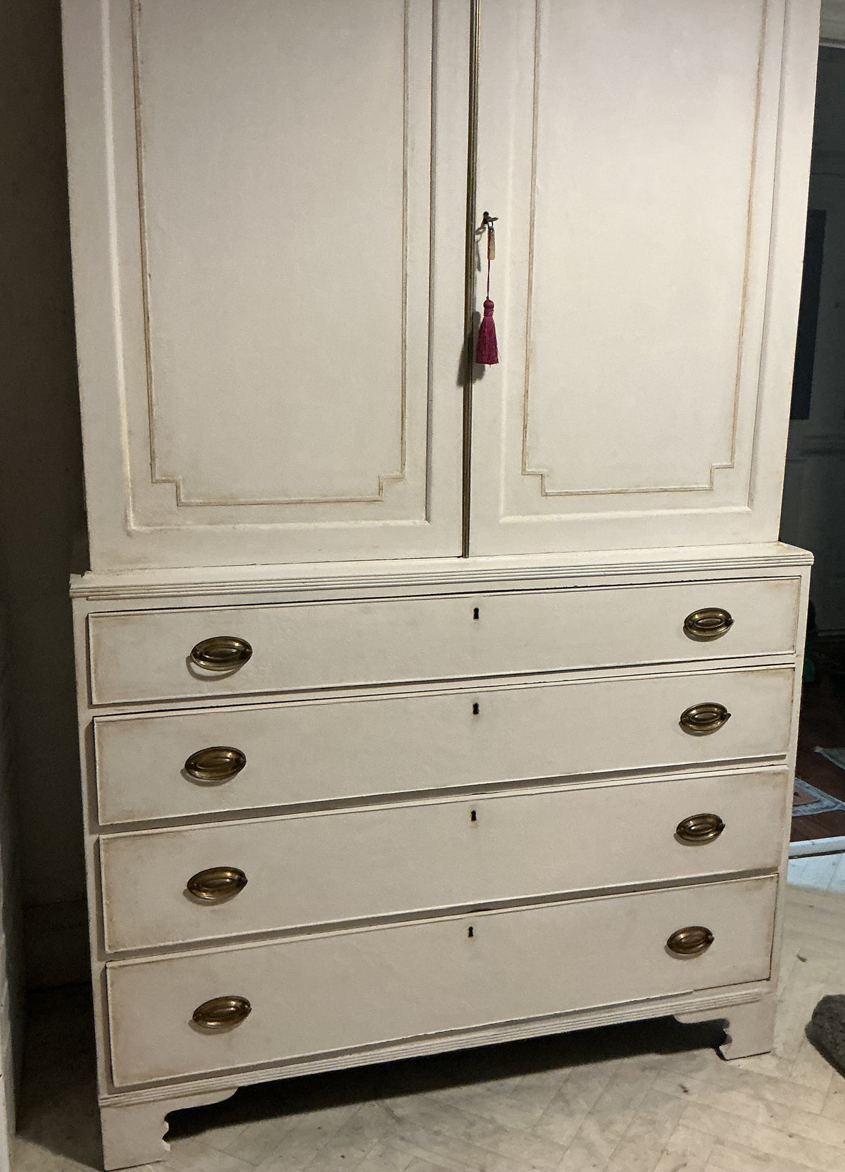 A tall, distressed white wooden cabinet with two upper doors and five drawers below, featuring brass oval handles and a small tassel keychain hanging from the center door handle