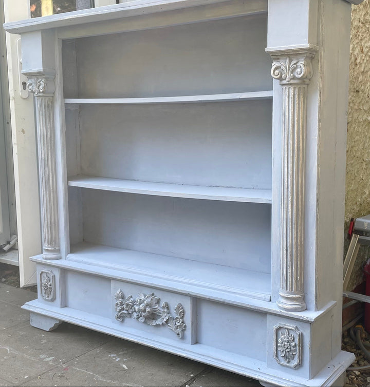 A tall, ornate three-tier bookcase with classical columns and decorative relief carvings, painted in a light gray finish, photographed outdoors on a patio.