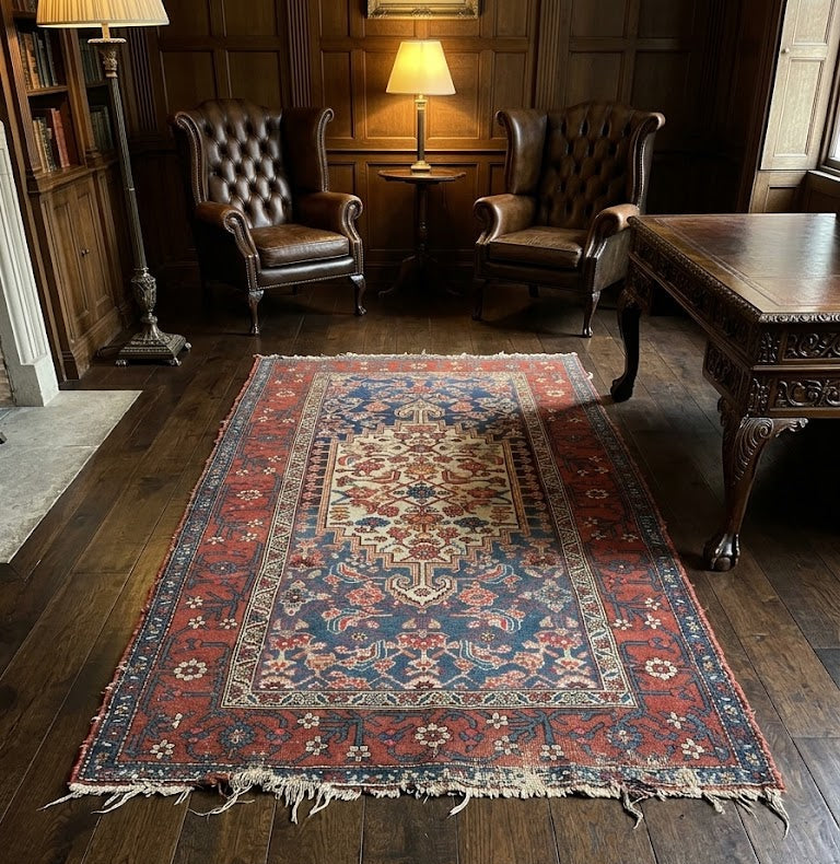traditional rectangular area rug with a red and blue floral pattern and cream fringe, displayed on a dark wood floor in a classic, dimly lit study room with leather armchairs and a wooden desk.