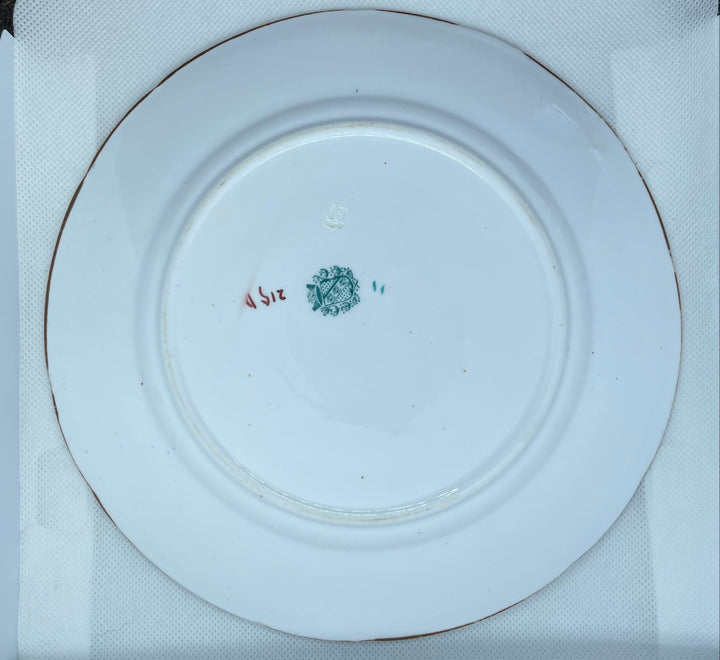 A white ceramic plate shown from the back, resting on a white textured mat. The plate features a small green and red mark in the center.
