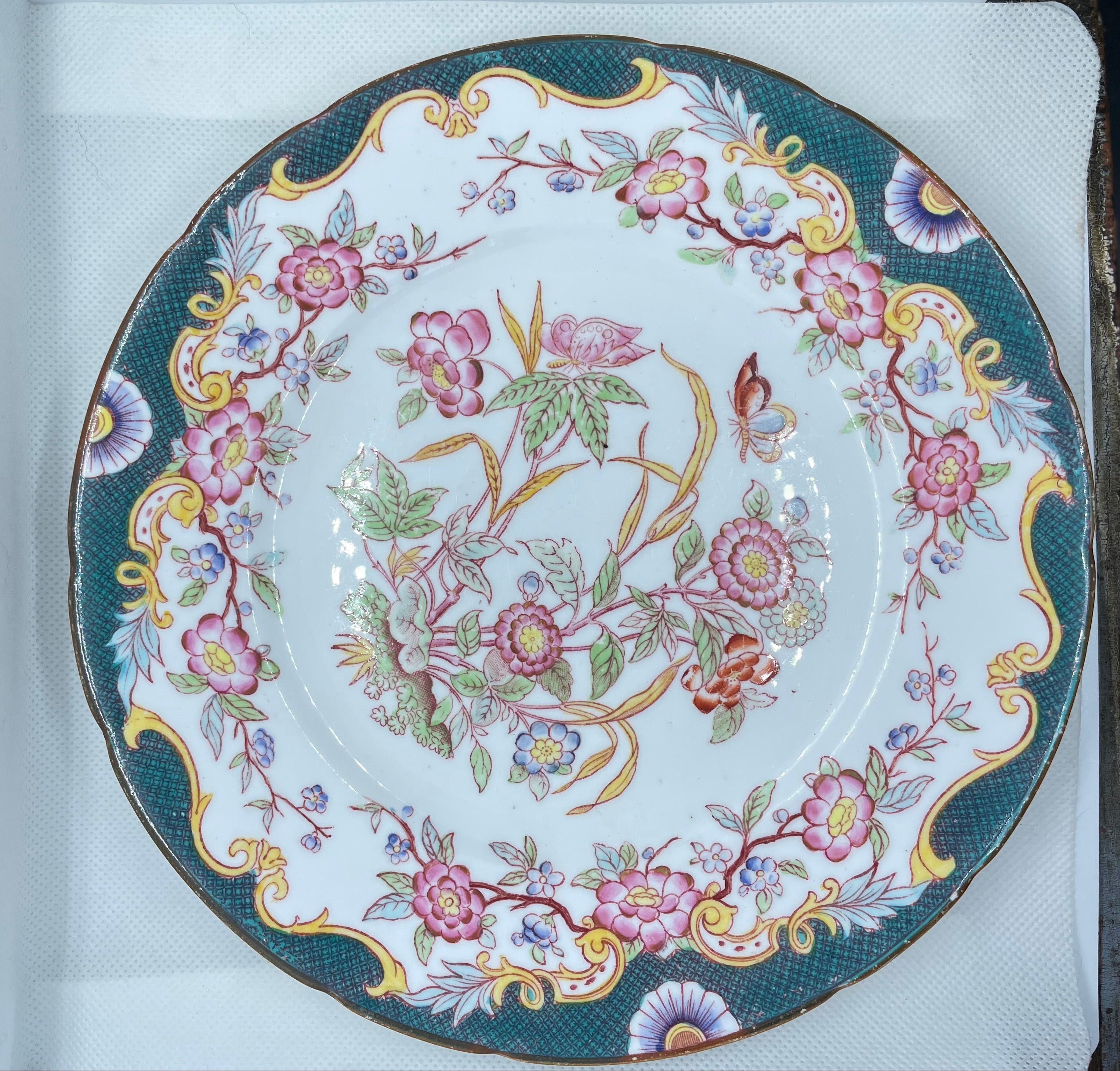 A round white porcelain plate with an ornate floral pattern, featuring pink and purple blossoms, green leaves, and gold accents on a dark teal border, displayed inside a white box.