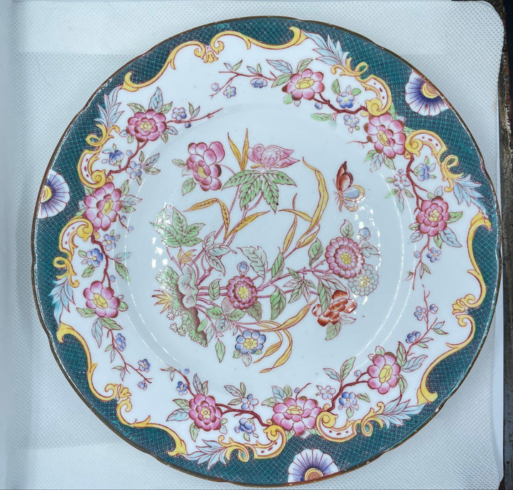 A round white porcelain plate with an ornate floral pattern, featuring pink and purple blossoms, green leaves, and gold accents on a dark teal border, displayed inside a white box.