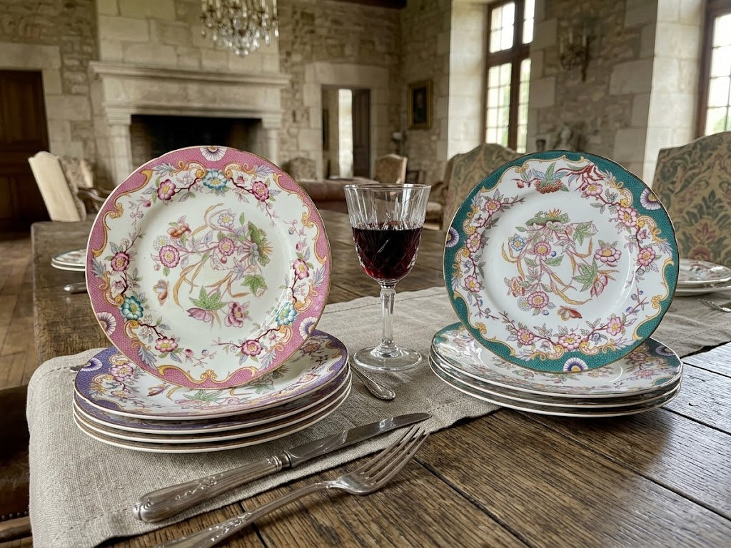 set of vintage floral porcelain plates displayed on a rustic wooden dining table in a stone room. The plates feature intricate hand-painted designs, including one with a pink border, one with a teal border, and others stacked underneath, alongside a glass of red wine and silver cutlery.