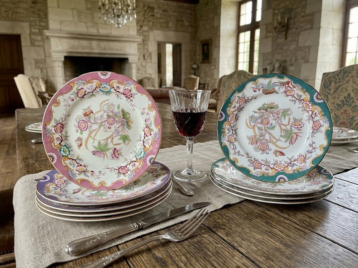 set of vintage floral porcelain plates displayed on a rustic wooden dining table in a stone room. The plates feature intricate hand-painted designs, including one with a pink border, one with a teal border, and others stacked underneath, alongside a glass of red wine and silver cutlery.