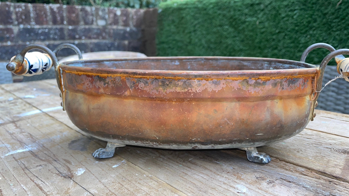 A rustic copper-colored metal bowl with decorative ceramic handles and small metal feet, displayed on a weathered wooden table with a garden background.