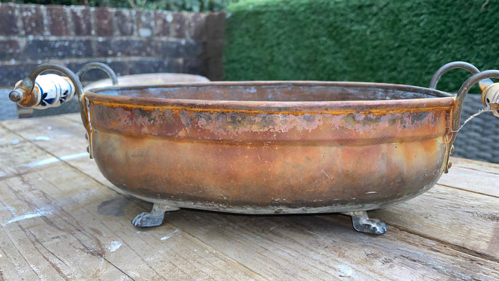A rustic copper-colored metal bowl with decorative ceramic handles and small metal feet, displayed on a weathered wooden table with a garden background.