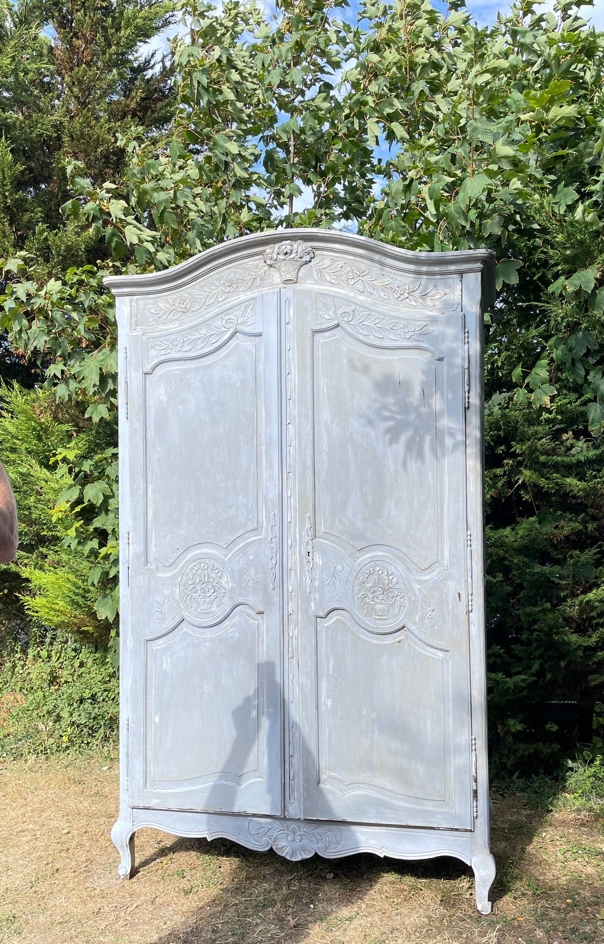 distressed white painted wooden wardrobe or armoire with ornate carved details, photographed outdoors on a grassy lawn with trees in the background.
