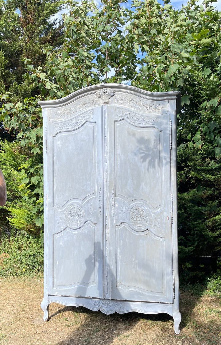 distressed white painted wooden wardrobe or armoire with ornate carved details, photographed outdoors on a grassy lawn with trees in the background.
