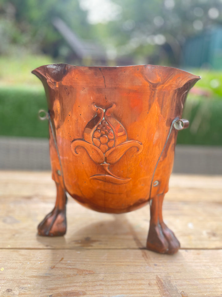 rustic carved wooden cup or planter with three legs, featuring a central floral relief and small metal rivets, set on a wooden surface with a blurred outdoor garden background.