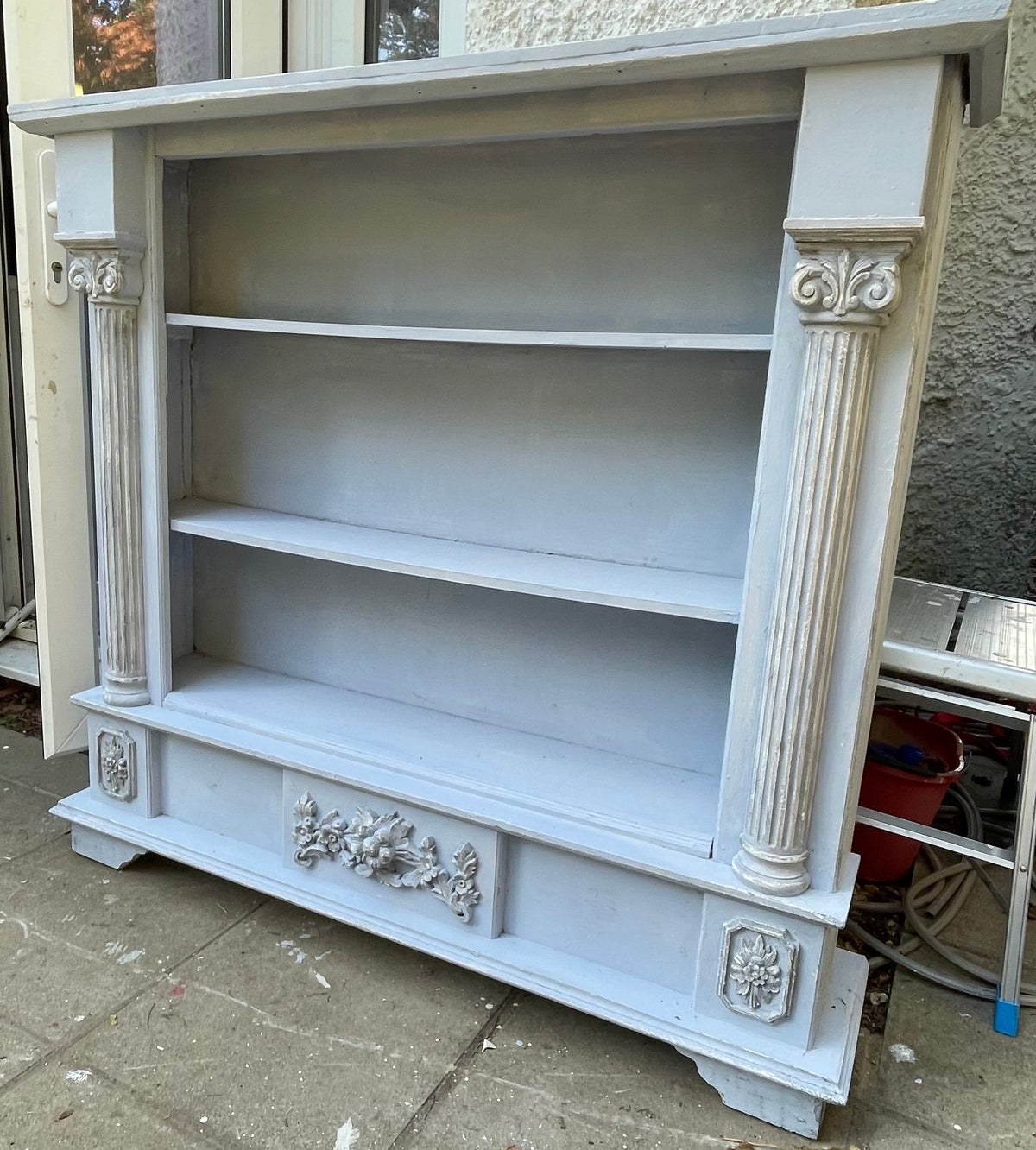  light gray painted wooden bookcase with three shelves and decorative carved details on the sides and bottom, photographed outdoors against a stucco wall and patio.